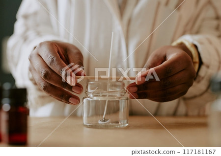 Hands crafting handmade candle in glass jar with wooden wick, surrounded by tools and ingredients for candle making. Process involves precise control and delicate handling 117181167