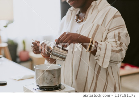 Person carefully pouring melted wax into a candle mold in a home studio setting, surrounded by equipment and tools essential for candle-making 117181172