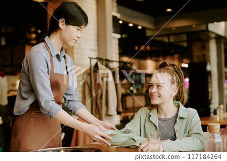 Asian waitress serving young Caucasian woman in cozy cafe with warm ambiance, creating friendly atmosphere and positive interaction between staff and customer 117184264