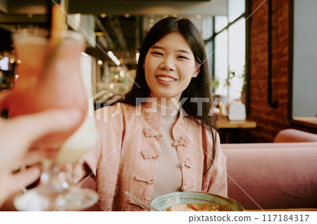 Asian woman smiling while raising glass in cozy restaurant setting, wearing traditional outfit and sitting at table with bowl of food 117184317