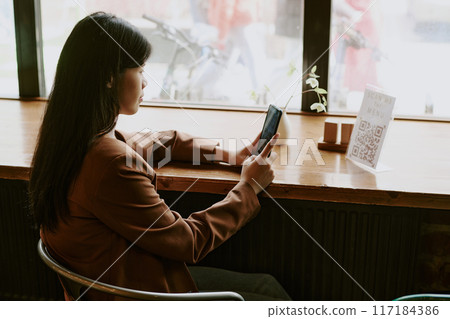 Asian woman sitting by window using smartphone and looking out at street view, creating ambiance of contemplation and productivity in quiet cafe setting 117184386