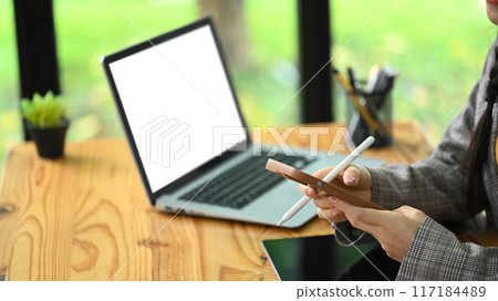 Young female office worker sitting at wooden desk with laptop and chatting on her smartphone 117184489