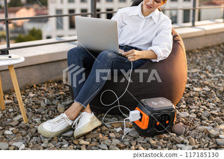 Business woman charging laptop with portable power station on rooftop 117185130