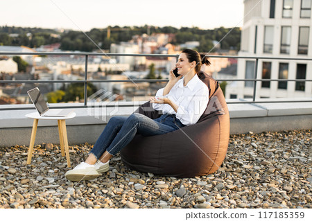 Woman working outdoors on rooftop with laptop and phone 117185359