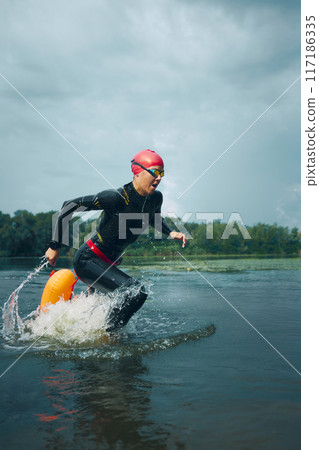 Athlete in wetsuit and swim cap splashes from lake with orange buoy attached, ready for rest amidst lush surroundings. 117186335