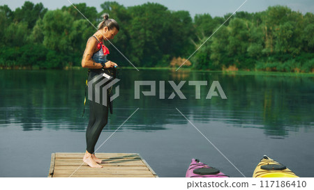 Female swimmer with warms up on dock by calm lake, surrounded by nature. Her wetsuit and gear indicate readiness for water sports. Female swimmer with warms up on dock by calm lake, surrounded by nature. Her wetsuit and gear indicate readiness for water sports. 117186410