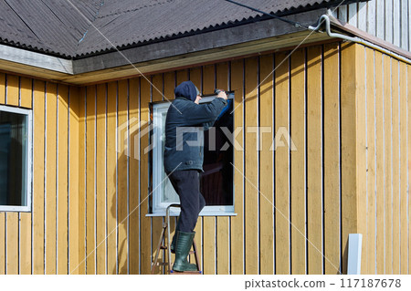 Roofing contractor does exterior trim of window opening while standing on steel step ladder in front of facade of wooden country house on an autumn afternoon. 117187678