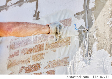 Brick cladding, also called brick veneer is attached to exterior of house made of aerated concrete blocks, construction worker uses tile adhesive. Brick cladding, also called brick veneer is attached to exterior of house made of aerated concrete blocks, construction worker uses tile adhesive. 117187712