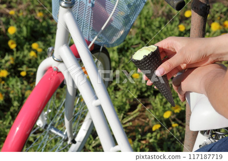 The girl is resting on the spring meadow. He sits on the trunk of the bike and eats ice cream. Dandelions are blooming, young grass is growing. 117187719