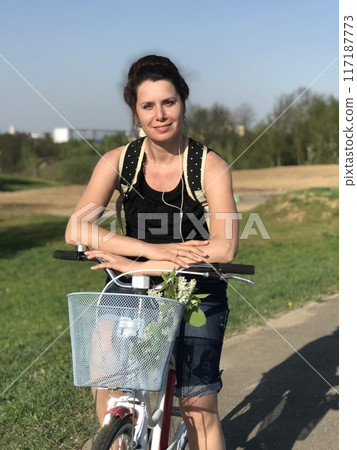 The girl is resting on the spring meadow. Sits on a bicycle, behind a tourist backpack. Dandelions are blooming, young grass is growing. The girl is resting on the spring meadow. Sits on a bicycle, behind a tourist backpack. Dandelions are blooming, young grass is growing. 117187773
