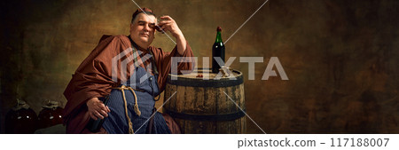 Hangover. Medieval man, monk in traditional attire leaning glass with beverage to head, leaning on wooden barrel with bottle Hangover. Medieval man, monk in traditional attire leaning glass with beverage to head, leaning on wooden barrel with bottle 117188007