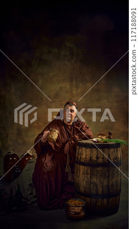 Man, medieval monk in monastic robes sitting in dark cellar, looking inside wooden mug of beer, leaning on barrels with onion and bread appetizers Man, medieval monk in monastic robes sitting in dark cellar, looking inside wooden mug of beer, leaning on barrels with onion and bread appetizers 117188091