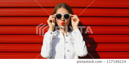 Stylish young woman blowing kiss in white glasses, fashionable female model posing on red background Stylish young woman blowing kiss in white glasses, fashionable female model posing on red background 117189224