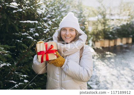 Happy young beautiful excited woman in white hat holding gift box in hands and smile on winter street. Winter holidays sale. Concept of shopping, holidays and happiness Christmas. High quality photo 117189912