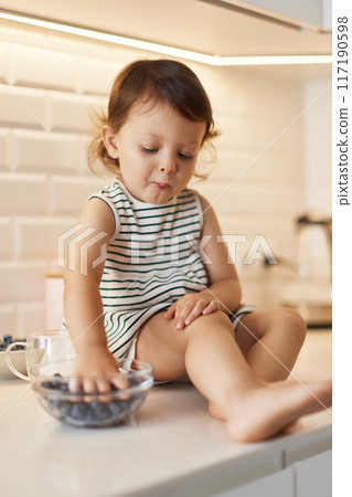 Little baby girl eating fresh blueberries from glass bowl in white kitchen 117190598