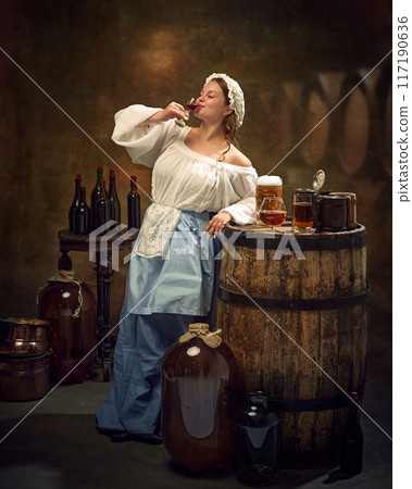 Woman in traditional attire drinking alcohol drink from glass, surrounded by barrels, bottles, and various brews at vintage pub. 117190636