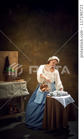 Young chubby woman, maiden, in historical dress, as waitress pours tea to cups on tray in vintage kitchenware. 117190723