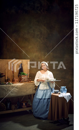 Young chubby woman, maiden, in historical dress holds tray, as waitress surrounded by vintage kitchenware and herbs. 117190725