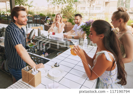 Young people, friends meeting in modern bar, open air restaurant, enjoying cocktails and conversation with male bartender 117190794