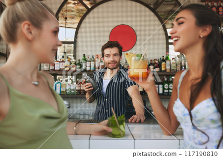 Focus on man, barman making delicious cocktails to two beautiful young women, meeting in bar and enjoying drinks and conversation Focus on man, barman making delicious cocktails to two beautiful young women, meeting in bar and enjoying drinks and conversation 117190810