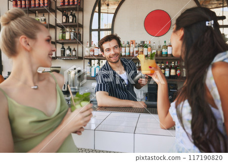 Two smiling young women meeting on modern outdoor bar, enjoying cocktails and cheerfully talking with man, bartender. Evening fun Two smiling young women meeting on modern outdoor bar, enjoying cocktails and cheerfully talking with man, bartender. Evening fun 117190820