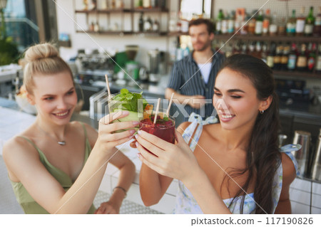 Cheers. Two smiling beautiful women clinking glasses with cocktails in modern bar settings, with bartender on background. Happiness and joy 117190826