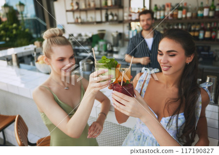 Cheers. Two smiling beautiful women clinking glasses with cocktails in modern bar settings, with bartender on background 117190827
