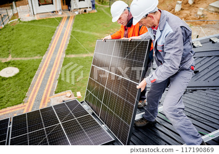 Roofers building solar panel system on roof of house. Men workers in helmets carrying photovoltaic solar module outdoors. Concept of alternative and renewable energy. 117190869