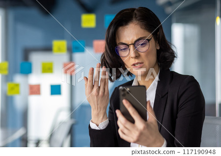 Businesswoman reading bad news on smartphone, looking worried in modern office. Woman wearing glasses, dressed in business attire, holding phone, distressed expression Businesswoman reading bad news on smartphone, looking worried in modern office. Woman wearing glasses, dressed in business attire, holding phone, distressed expression 117190948