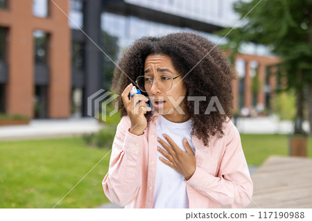 African American woman with using asthma inhaler outdoors. Young woman looking concerned or anxious while using inhaler to manage asthma symptoms. Concept of asthma, health, and respiratory issues African American woman with using asthma inhaler outdoors. Young woman looking concerned or anxious while using inhaler to manage asthma symptoms. Concept of asthma, health, and respiratory issues 117190988