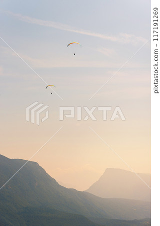 Two paragliders in the air fly over the mountains at summer sunrise. Two paragliders in the air fly over the mountains at summer sunrise. 117191269