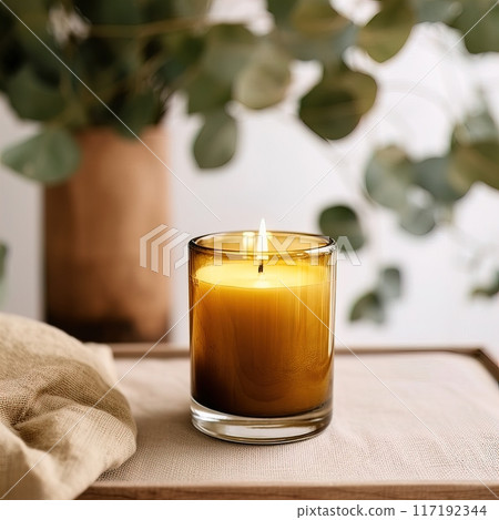 A lit candle in an amber glass jar against fresh eucalyptus leaves creates a calming ambiance 117192344