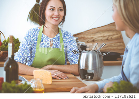 Two women friends taste new recipes for delicious fondue while sitting at the kitchen table. Healthy meal cooking concept Two women friends taste new recipes for delicious fondue while sitting at the kitchen table. Healthy meal cooking concept 117192621