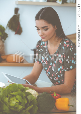 An attractive young dark-haired woman chooses the recipe for a delicious meal while sitting at the table in the kitchen. Tablet pc is the best cookbook 117192713