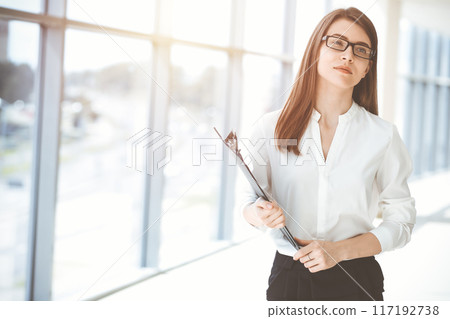 Beautiful female specialist with clipboard standing in sunny office and smiling charmingly. Business people concept Beautiful female specialist with clipboard standing in sunny office and smiling charmingly. Business people concept 117192738