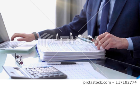 Businessman using a calculator and magnifying glass while analyzing financial documents at desk in fair modern office. Audit and taxes in business Businessman using a calculator and magnifying glass while analyzing financial documents at desk in fair modern office. Audit and taxes in business 117192774