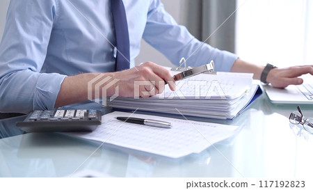 Businessman in blue short is using calculator and reviewing ring folder of financial documents with magnifying glass at desk in modern office. Audit and taxes in business concept 117192823