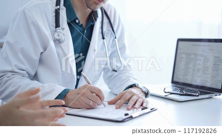 Doctor and a patient. The physician, wearing a white medical coat over a green shirt, is filling out a medical record form during a consultation in the clinic. Medical service 117192826