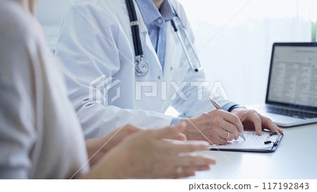 A doctor and a patient. The physician, wearing a white medical coat over a blue shirt, is filling out a medical record form during a consultation in the clinic. Medical service A doctor and a patient. The physician, wearing a white medical coat over a blue shirt, is filling out a medical record form during a consultation in the clinic. Medical service 117192843