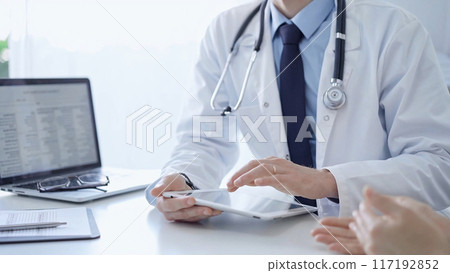 Doctor and a patient. The physician, wearing a white medical coat over a blue shirt and tie, is using tablet computer during a consultation in the clinic. Medicine 117192852