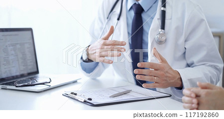 Doctor and a patient. The physician, wearing a white medical coat over a blue shirt and tie, gesturing with his hands during a consultation in the clinic. Medicine concept Doctor and a patient. The physician, wearing a white medical coat over a blue shirt and tie, gesturing with his hands during a consultation in the clinic. Medicine concept 117192860