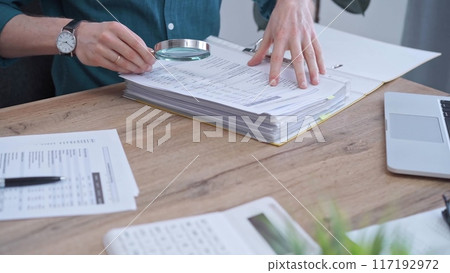 Businessman or auditor wearing green shirt is reviewing financial documents with magnifying glass. Business work process in bookkeeping office. Taxes 117192972