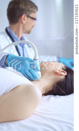 Doctor man wearing blue protective gloves is using ultrasound equipment on the neck of a patient woman, lying for examination. Medicine concept 117193021