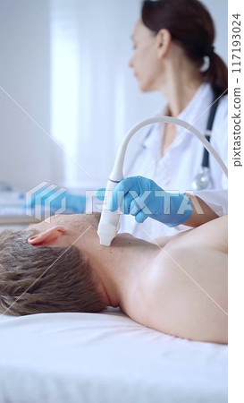 Doctor woman wearing blue medical gloves performing neck ultrasound exam on male patient. Medicine service concept Doctor woman wearing blue medical gloves performing neck ultrasound exam on male patient. Medicine service concept 117193024