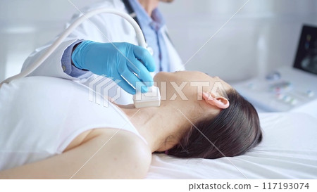 Doctor man wearing blue medical gloves is using ultrasound equipment on neck of female patient lying down for a medical examination 117193074