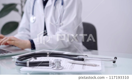 Clipboard with medical records and stethoscope are lying on the glass table while doctor woman is using tablet computer on the glass table. Medicine concept 117193310
