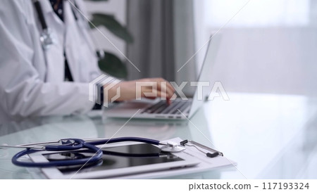Tablet computer and blue stethoscope are on the glass table while doctor woman is using laptop computer on the glass table. Medicine concept Tablet computer and blue stethoscope are on the glass table while doctor woman is using laptop computer on the glass table. Medicine concept 117193324
