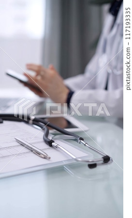 Stethoscope and medication history record form are on the glass table while doctor woman is using smartphone. Medicine theme 117193335
