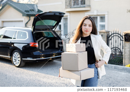 Young business woman picking up parcels from a car trunk, coming home by car. Concept of buying goods online and delivering them home 117193468