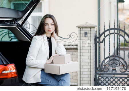 Girl sitting in the trunk of the car, holding boxes in his hands and smiling, looking at the camera. Concept of buying goods online and delivering them home 117193478